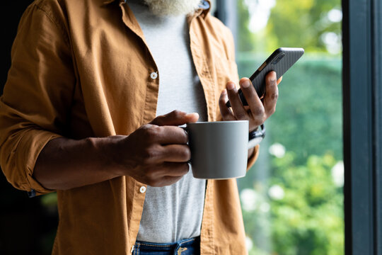 Midsection of senior african american man holding coffee mug using smartphone at home