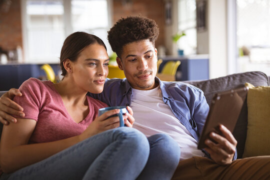 Young biracial couple watching video together on digital tablet sitting on sofa at home