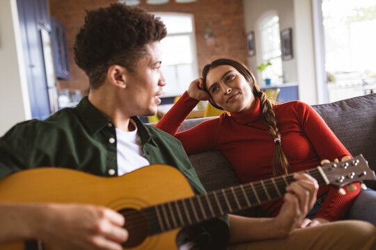 Young biracial man playing acoustic guitar and singing for girlfriend sitting on sofa in living room