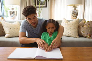 Biracial father helping blind daughter reading book while touching braille at home