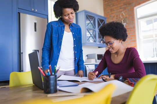 Mother looking at daughter writing in book during online lecture on laptop at home - Powered by Adobe