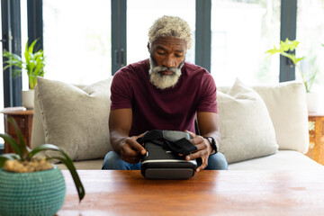 Bearded african american senior man holding virtual reality headset sitting on sofa in living room