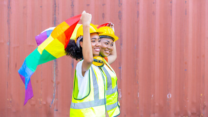 Young female African sisters in safety uniform and helmet show LGBT flag to support freedom and rights for gender equality next to red cargo or shipping container