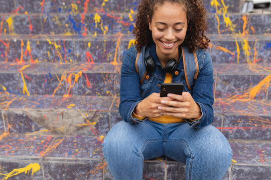 Smiling young african american woman using smartphone while sitting on steps with graffiti