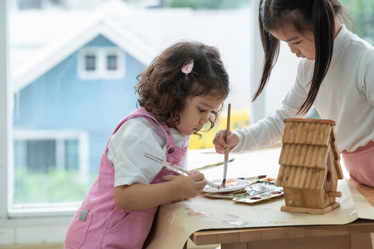 Two little girls help paint a wooden birdhouse in the room, education and learning concept