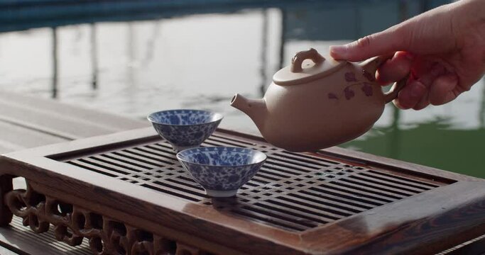Beautiful Chinese tea ceremony near water pool slow motion eastern culture style. Female hand pouring fresh steaming drink into cups. Meditative calm mood and atmosphere