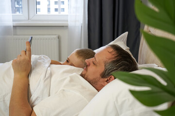 Obraz premium Father and son are watching funny video on phone in bed and smiling