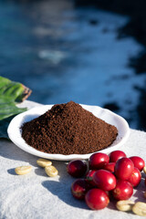 Red ripe arabica coffee berries, green coffee beands, leaves and roasted ground coffee in bowl and blue ocean view