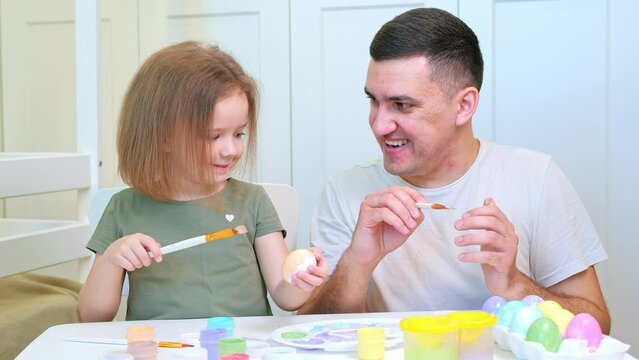 A young dad with a short haircut and his little daughter are sitting at a white drawing table. Needlework, do it yourself, preparation for Easter holiday.
