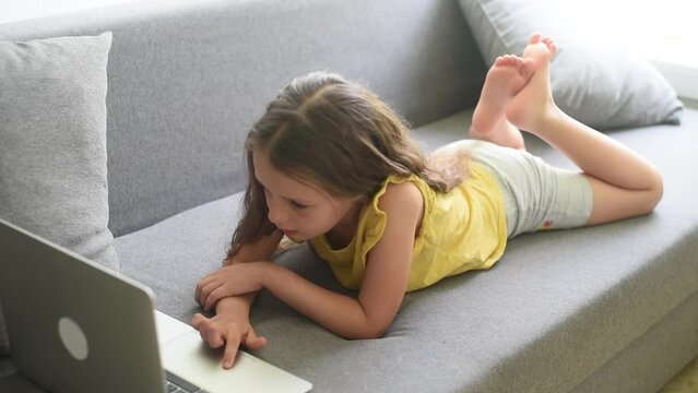Cute child girl using laptop, lies on couch, little smart kid typing on computer chatting with friend, talk online, study at home, playing and watch cartoons on gadget.