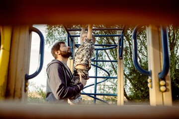 a boy doing chin-ups on monkey bars and father is helping him. Exercises on the playground on the beach. father's day concept