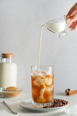 Refreshing iced coffee with milk topping pouring from gravy boat in drinking glass with ice cubes served on plate with roasted beans on white wooden table with brown sugar, metal straw and jar