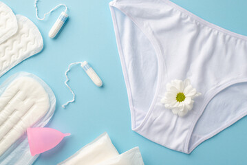 Top view photo of white cotton classic lingerie with camomile flower bud sanitary napkins menstrual cup and tampons on isolated pastel blue background