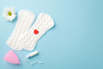 Top view photo of two panty liners with red heart camomile bud tampon and pink menstrual cup on isolated pastel blue background with copyspace