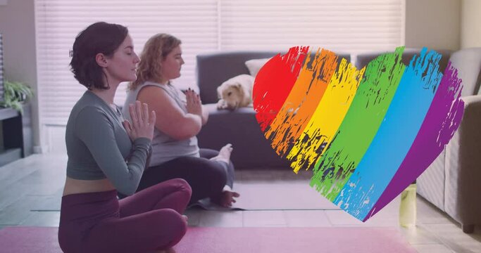 Animation of rainbow heart over caucasian lesbian couple practicing yoga at home