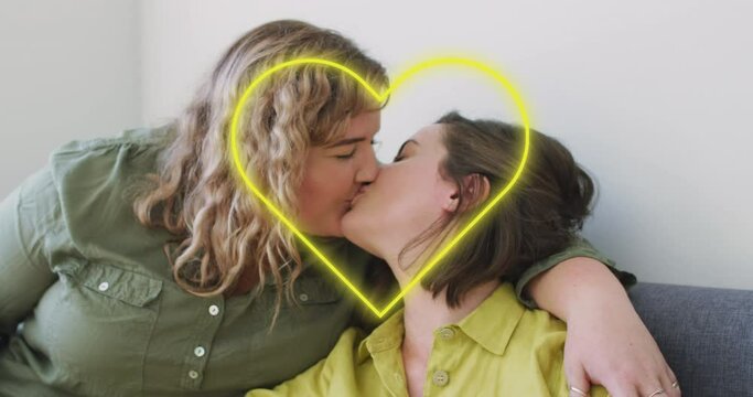 Animation of neon heart over happy caucasian lesbian couple kissing at home