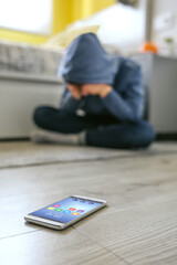 Unrecognizable teenager boy crying desperate for bullying with cell phone lying on the floor in foreground. Selective focus on mobile in foreground