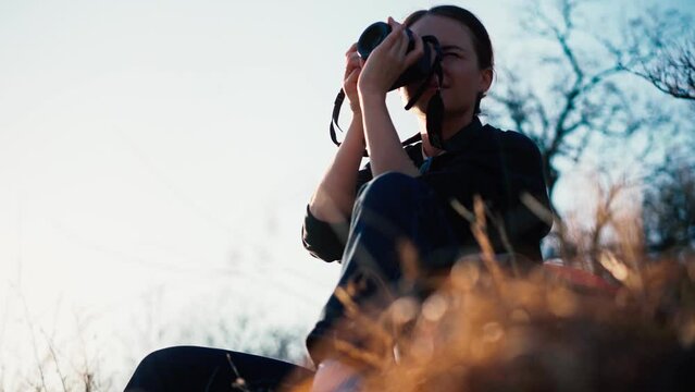 A young beautiful woman traveler taking a picture with her camera and enjoying the view while sitting on the hill at sunset time.