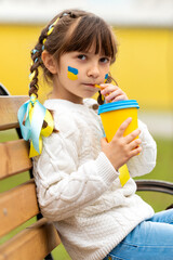 A little Ukrainian girl with two pigtails and yellow and blue ribbons drinks a hot drink from a glass of patriotic colors like the Ukrainian flag. Stop the war in Ukraine. Volunteer Help for Refugees