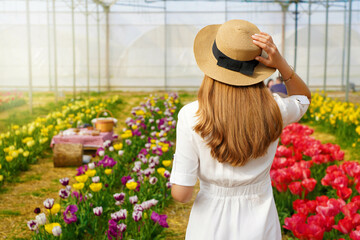 Beautiful girl holds straw hat walking between flowers ready for picnic on sunset. Back view.