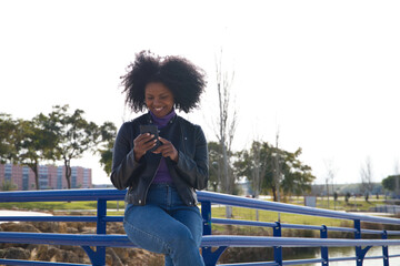 Obraz premium Young, beautiful Afro-American woman with mobile phone shopping online, checking her email and social networks in the park. The woman is wearing casual and comfortable clothes and is happy.