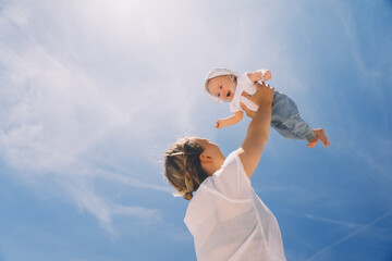 Mother throwing baby up against the blue sky. Happy family outdoors. Mom and baby at summer on...