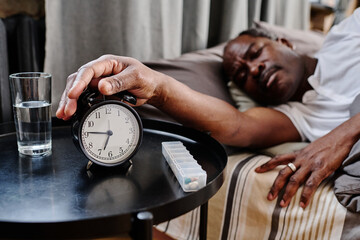 Hand of senior African American sleepy man pressing buzz bell of alarm clock on small round table...