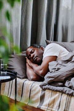 Tired Mature African American Man Sleeping Peacefully In Bed In The Morning In Front Of Small Black Table With Glass Of Water