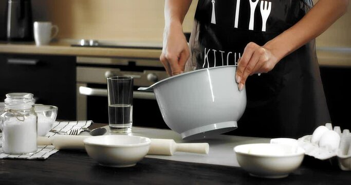 Young brunette woman in a white T-shirt and a brown apron is standing at a table in a modern kitchen, stirring dough in a gray bowl with a whisk to bake shortbread cookies. Baking Ingredients.Upward