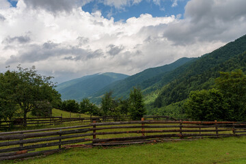 Empty pasture in the mountains fenced with a wooden fence, against the backdrop of the forest, sky and clouds