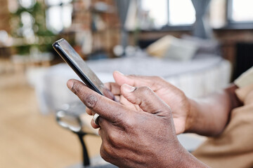 Hands of contemporary retired black man holding mobile phone and texting at free time in his bedroom during everyday routine