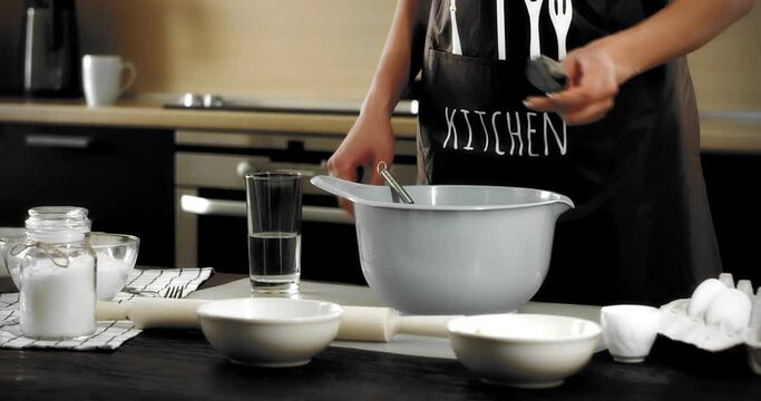 Young woman in a brown apron stands at a table in a modern kitchen, spooning flour into a gray bowl to bake shortbread cookies. Baking Ingredients