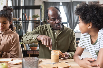 Contemporary senior African American man making sandwich for his grandson during breakfast by served table in the morning
