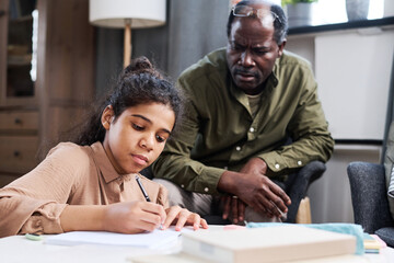 Serious multi-ethnic schoolgirl making notes in copybook while doing homework and her grandfather...