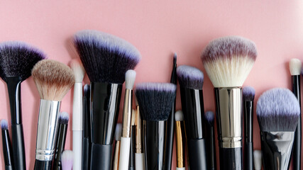 many different makeup brushes lie from below on a pink background