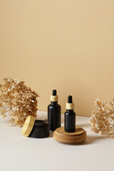 Black dropper cosmetic bottles and jar of moisturizer cream with dry flowers on beige background.