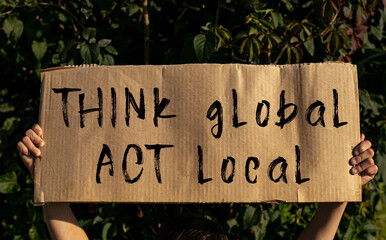 A poster with the words THINK GLOBAL, ACT LOCAL in the hands of a young girl on a background of green foliage.