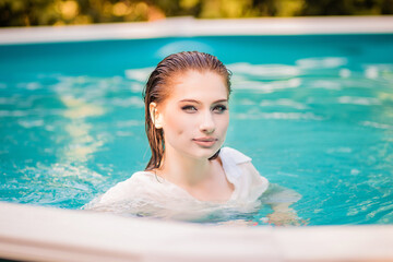 Portrait of a beautiful girl with wet hair floating in the pool, wearing a white shirt. Rest in the water in hot weather.
