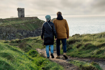 Couple walking holding hands on a foot path of Cliff of Moher, Ireland. Moher Tower at Hag's Head in the background. Travel and tourism concept. Exploring stunning nature scene. 