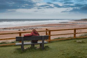 Fototapeta na wymiar Teenager sitting on a bench looking at beautiful Fanore beach. Ireland. Blue cloudy sky and Atlantic ocean. Travel and tourism concept.