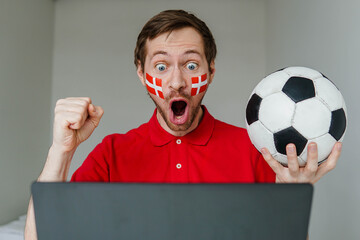 Young excited man fan support Denmark national football team hold in hand soccer ball watch tv live stream on laptop.
