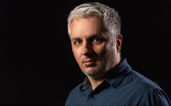 Head and shoulders low key portrait of a bearded middle-aged man looking thoughtfully at the camera over a black studio background with copy space