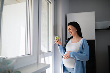 Pregnant caucasian in good mood looks at fresh green apple that is in her right hand and stands...