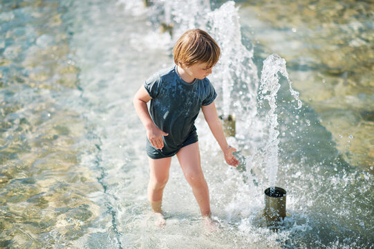 Outdoor portrait of happy little boy playing inside of city fountain on a hot summer day