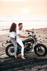 Fototapeta na wymiar male and female couple in white outfit sitting on a motorcycle at a black sand beach in Bali Indonesia on a black vintage motorcycle during sunset
