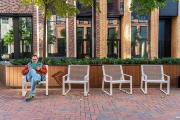 Millennial man with happy face checking phone sitting on one of four chairs in courtyard of modern  residential complex with landscaping. Portrait of excited man holding smart phone at street outdoors