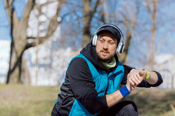 Modern technology and active lifestyle. A smiling athlete exercises with a fitness bracelet, listens to music on headphones, rests on a bench after a run.