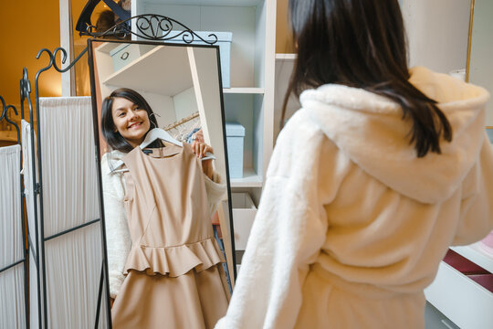 Smiling young woman in a bathrobe choosing a dress in front of a mirror at home or in a hotel
