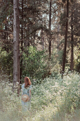 Portrait of a dreamy and relaxed happy red-haired girl in a linen rustic dress and a straw hat in a pine grove surrounded by white wildflowers. Walking in forest