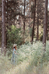 Portrait of a dreamy and relaxed happy red-haired girl in a linen rustic dress and a straw hat in a pine grove surrounded by white wildflowers. Walking in forest